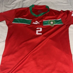 Marocko fotbollströja Hakimi #2 Puma - Säljer en röd Marocko landslagströja från Puma med gröna och vita detaljer, nummer 2 och Hakimi på ryggen. Tröjan har korta ärmar, grönt kragparti och broderat landslagsemblem på bröstet. Perfekt för fotbollsfans!
