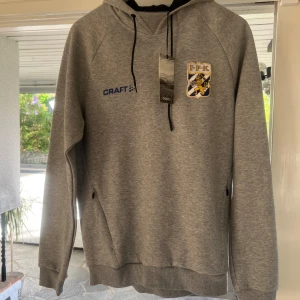Grå hoodie från Craft med IFK-emblem - Säljer en grå hoodie från Craft med IFK-emblem på bröstet. Tröjan har huva med svarta snören, ribbade muddar och fickor med dragkedja. Perfekt för dig som vill visa ditt stöd för IFK! Storlek M