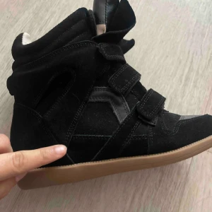 Svarta sneakers med kilklack från Isabel Marant - Säljer ett par svarta sneakers med kilklack från Isabel Marant. Skorna har tre kardborreband, är gjorda i mocka och skinn, och har en beige sula. De har en sportig och trendig design med högre skaft.