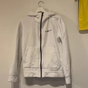 Vit hoodie från Sail Racing - Vit hoodie från Sail Racing med svart dragkedja och logga på bröstet. Tröjan har huva och två stora fickor framtill. Perfekt för en sportig och avslappnad stil. Finns även en i svart!!!