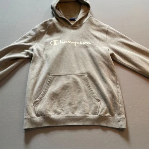 Grå hoodie från Champion - Säljer en grå hoodie från Champion med klassisk logga framtill. Tröjan har huva, magficka och långa ärmar. Perfekt för en avslappnad stil och enkel att matcha med det mesta. Står XL men sitter som L
