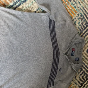 Grå stickad tröja med dragkedja från Chaps - Säljer en grå stickad tröja från Chaps med halv dragkedja och ribbad krage. Tröjan har mörkgrå ränder över bröstet och ett broderat emblem på vänster sida. Perfekt för kyliga dagar.