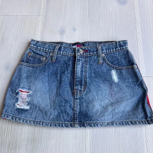 Jeanskjol med slitningar och röda detaljer - Säljer en blå jeanskjol med slitna detaljer och röda sömmar. Kjolen har klassiska fickor fram och bak samt bälteshällor. Kort modell med råa kanter och en cool, avslappnad look.