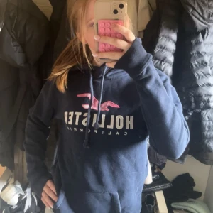Mörkblå hoodie från Hollister - Säljer en jättefin mörkblå hoodie från Hollister med vit och rosa logga på bröstet. 