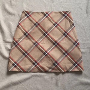 Beige rutig kjol från H&M - Snygg beige kortkjol från H&M med klassiskt rutigt mönster i rött, svart och vitt. Kjolen har en rak passform och är fodrad på insidan för extra komfort. Perfekt att styla med en enkel topp eller stickad tröja.