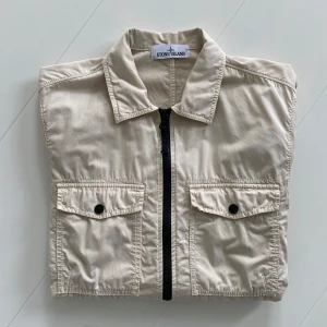 Beige Overshirt från Stone Island - Snygg beige Overshirt / jacka från Stone Island med klassisk krage och svarta detaljer. Jackan har två bröstfickor med lock och knappar, dragkedja framtill och den ikoniska Stone Island-loggan på ärmen. Köpt i butik i Stockholm.