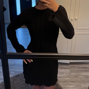 Svart plisserad klänning med långa ärmar - Säljer en svart kortklänning med plisserat tyg och långa ärmar. Klänningen har en rund halsringning och lätt utsvängda ärmslut för en trendig look. Perfekt för dig som gillar stilrena och moderna plagg.