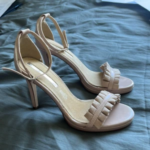 Beige sandaler med volangdetalj och klack - Säljer ett par stilrena rosa/beige klackskor med volangdetalj framtill. Skorna har öppen tå och smal rem runt vristen för extra stöd. Perfekta nu inför balen😌!! Jag säljer pgr av att dem är för stora för mig då jag fick dem av en kommpis.