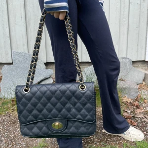 Svart quiltad axelväska med kedjerem - Säljer en svart handväska med snygg kedjerem i guld och svart. Väskan har ett rutmönster och ett vridlås i guld framtill. Perfekt storlek för det viktigaste och ger en stilren look. Får plats med det mesta i den💘