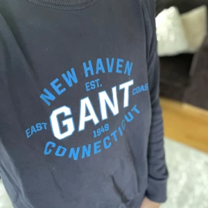 Mörkblå sweatshirt från GANT - Säljer en mörkblå sweatshirt från GANT med tryck i blått och vitt på bröstet: 'NEW HAVEN GANT CONNECTICUT'. Tröjan har rund halsringning och långa ärmar. Perfekt för en avslappnad stil.