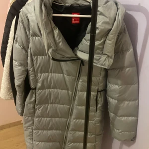 Ljusgrå dunjacka från Nike - Säljer en ljusgrå dunjacka från Nike med huva och dragkedja. Jackan är quiltad och har lång ärm, perfekt för kyliga dagar. Enkel och stilren design som passar till många olika outfits.
