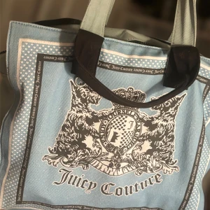 Blå axelväska från Juicy Couture - Säljer en blå axelväska från Juicy Couture med stort tryck och emblem framtill. Väskan har svarta handtag och detaljer samt innerfoder med Juicy Couture-logga. Perfekt för att bära med sig det viktigaste. PRIS KAN DISKUTERAS!
