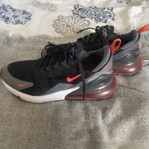 Svarta Nike Air Max 270 sneakers med röda detaljer - Nike Air Max 270 sneakers i svart med grå och röda detaljer. Skorna har en stor synlig Air Max-dämpning i hälen, mesh-överdel och snörning. Snygg och sportig design med vit sula och rund tå.