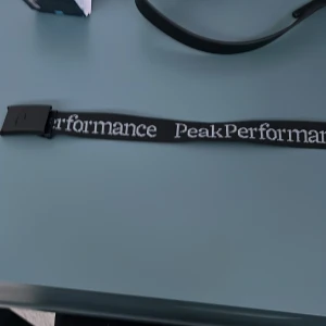 Svart bälte från Peak Performance - Säljer ett svart bälte från Peak Performance med tydlig logga längs bandet. Bältet har en svart spänne i metall och är gjort i ett slitstarkt tygmaterial. Perfekt accessoar för att lyfta din outfit.
