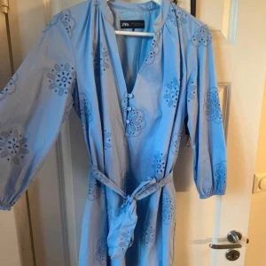 Ljusblå klänning med broderade blommor från Zara - Söt ljusblå klänning från Zara med broderade blommor och knappar framtill. Klänningen har trekvartsärm, v-ringning och ett matchande knytbälte i midjan. Perfekt för vår och sommar!