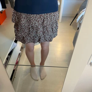 Brun blommig volangkjol från Lindex - Söt brun kortkjol från Lindex med vitt blommönster och flera volanglager. Kjolen har bred resår i midjan för extra komfort och en luftig känsla. Perfekt för varma dagar och enkel att matcha med olika toppar. kjolen är i storlek 134-140, men skulle säga att den passar XXS,XS och S. 