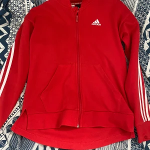 Röd zip-hoodie från Adidas - Röd hoodie med dragkedja från Adidas. Klassiska vita ränder längs ärmarna och Adidas-logga på bröstet. Två fickor framtill och huva. Knappt använd! Strl 164/14Y. 50kr