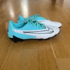 Nike Phantom fotbollsskor i blått och vitt - Säljer ett par Nike Phantom fotbollsskor i fräscha färger – ljusblå och vit med svarta detaljer. Den vänstra skon har en liten defekt på Nike märket fast annars är skon i riktig bra skick. Pris kan diskuteras vid snabb affär. 