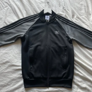 Svart och grå Adidas track jacket - Säljer en klassisk svart och grå Adidas track jacket med dragkedja och en grå Adidas-logga på bröstet. Perfekt för en sportig och avslappnad stil.😮‍💨Säljer så min bror inte använder den längre, precis som ny🤩‼️