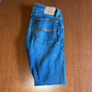 Blå jeans från Nudie Jeans - Säljer ett par klassiska blå jeans från Nudie Jeans med orange sömmar och broderad bakficka. Jeansen har normal passform. Perfekta för dig som gillar stilrena och tidlösa jeans. W26 men passar mig som har W29-W31 i midjan för dom är lite stretchy 
