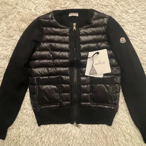 Svart pufferjacka från Moncler - Säljer en svart pufferjacka från Moncler med quiltad framsida och ärmar i stickad bomull. Jackan har dragkedja framtill och rund hals. Klassisk Moncler-logga på ärmen och två fickor framtill.