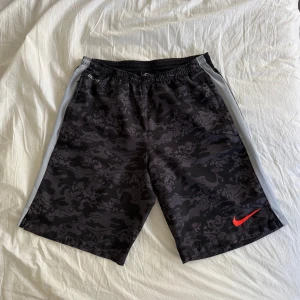 Svarta camo shorts från Nike - Säljer ett par svarta shorts från Nike med grått camouflage-mönster och röd swoosh-logga. De har elastisk midja och vita sidopaneler. Perfekta för träning eller chill.