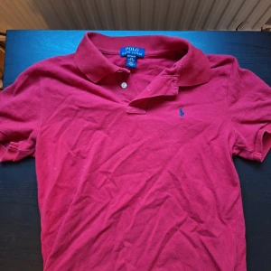 Röd pikétröja från Polo Ralph Lauren - Säljer en klassisk röd pikétröja från Polo Ralph Lauren med blå broderad logga på bröstet. Tröjan har krage, knappar vid halsen och är tillverkad i bomull. Perfekt för en stilren och avslappnad look.