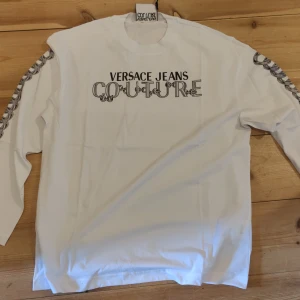 Vit långärmad tröja från Versace Jeans Couture - Vit långärmad tröja med tryckt Versace Jeans Couture-logga framtill och kedjedetaljer längs ärmarna. Rund halsringning och klassisk passform. Perfekt för dig som gillar stilrena plagg med exklusiv känsla.