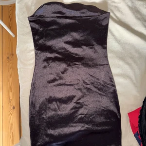 Svart axelbandslös satinklänning - Säljer en vinröd axelbandslös klänning i glansigt satinmaterial. Klänningen är figurnära och har en kort längd, perfekt för dig som vill ha en stilren och elegant look. Passar till många olika tillfällen!
