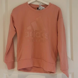 Rosa sweatshirt från Adidas - Säljer en rosa sweatshirt från Adidas med diskret logga och text framtill. Tröjan har rund halsringning och långa ärmar. Perfekt för en avslappnad stil.