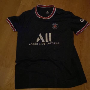 PSG fotbollströja Messi 30 - Säljer en svart PSG fotbollströja med Messi och nummer 30 på ryggen. Tröjan har korta ärmar, röd-vit-blå detaljer på krage och ärmslut samt klubbmärke och sponsortryck på bröstet.