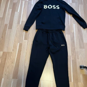 Svart hoodie och byxor från BOSS - Säljer ett svart set från BOSS med matchande hoodie och byxor. Hoodien har guldigt BOSS-tryck på bröstet, huva med dragsko och magficka. Byxorna har samma guldiga BOSS-tryck på ena benet och dragsko i midjan. Jätte fint skick inte blekt el nåt . Hoodie strl XS byxor S 