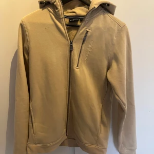 Beige ziphoodie från Sail Racing - Säljer en beige ziphoodie från Sail Racing med dragkedja framtill och en bröstficka med dragkedja. Jackan har huva och diskret logga på bröstet. Perfekt för dig som gillar stilren och sportig design.