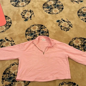 Ljusrosa croppad tröja med halv dragkedja - Säljer en ljusrosa långärmad tröja med halv dragkedja framtill och hög krage. Modellen är croppad och har en avslappnad passform. Perfekt för en chill och stilren look.