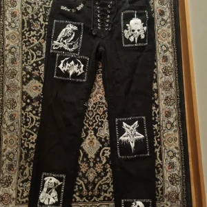 Svarta byxor med patchar och snörning från Widow - Unika svarta byxor från Widow med flera vita patchar med metal/goth-motiv och snörning framtill i midjan. Byxorna är storlek S men passar mer xs/s och från Widow. 