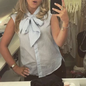 Blårandig ärmlös blus med rosett - Säljer en söt blårandig ärmlös blus med stor rosett framtill. Perfekt för en fräsch och somrig look. 