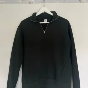 Mörkgrön halvzip tröja från Zara - Säljer en mörkgrön långärmad tröja från Zara med halv dragkedja framtill och hög krage. Tröjan har en enkel och stilren design, perfekt för lager-på-lager. Mjuk insida och ribbade muddar vid ärmslut och nederkant.