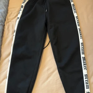 Svarta mjukisbyxor med vita sidor från Mainfinity - Svarta mjukisbyxor från Mainfinity med vita sidor där det står 'DREAM' i svart text. Byxorna har dragsko i midjan och är perfekta för en avslappnad stil. Mjukt material och bekväm passform.