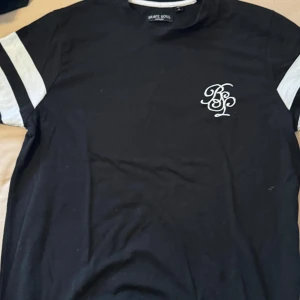 Svart t-shirt från Brave Soul med vita detaljer och shorts  - Svart t-shirt från Brave Soul med vita ränder på ärmarna och vit BS-logga på bröstet. Klassisk passform och rund halsringning. Perfekt för en avslappnad stil med shorts 