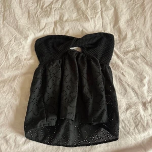Svart tubtopp  - Säljer en svart tubtopp med snygg detalj framtill. Toppen har broderade blommönster och är tillverkad i ett lätt, luftigt material. Perfekt för varma dagar eller när du vill ha en extra fin look.