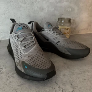 Grå Nike Air Max 270 sneakers - Snygga grå Nike Air Max 270 sneakers med svarta och blå detaljer. Skorna har ventilerande mesh-ovandel, snörning och en tydlig Air Max-sula för extra komfort. Perfekta för vardagsstil och träning.