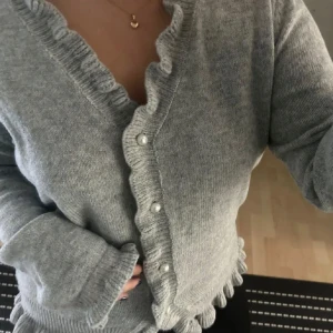 Grå kofta med volangkant och pärlknappar - Superfin grå kofta med volangkant längs v-ringningen och nederkanten. Stängs med pärlknappar framtill. Mjuk och stickad, perfekt för lager-på-lager. Snygg detalj med volanger som ger en söt touch.