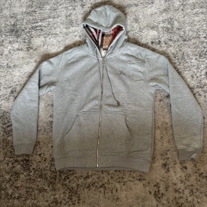 Grå hoodie från Burberry med dragkedja - Säljer en grå hoodie från Burberry med dragkedja och rutigt foder i huvan och på insidan. Tröjan har två fickor framtill och broderad logga på bröstet. 