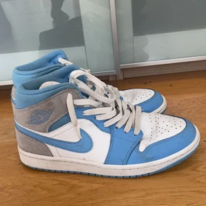 Jordan 1 Mid University Blue Gray  - Säljer ett par Nike Air Jordans med vit bas, ljusblå detaljer och grå mocka vid hälen. Skorna har snörning och rund tå, samt klassisk Air Jordan-logga på sidan. Perfekta för dig som gillar streetwear och sneakers med stil.