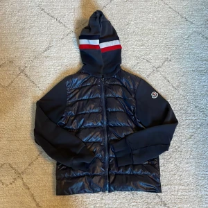 Moncler cardigan  - Säljer en snygg svart jacka från Moncler med huva och dragkedja. Jackan har ett coolt randigt mönster i rött och vitt på huvan och ett Moncler-märke på ärmen. Perfekt för kyligare dagar! Köptes begagnat men passade inte mig så säljer om! 