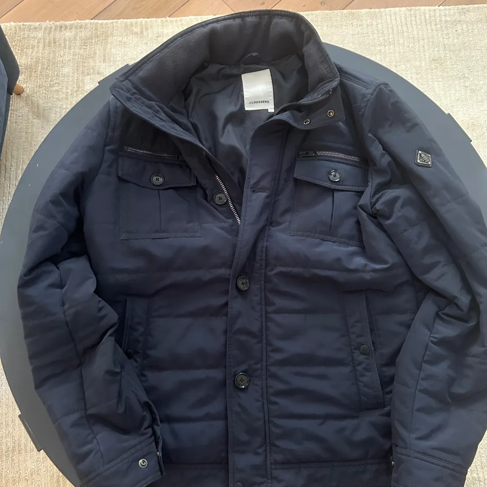 Säljer en stilren mörkblå field jacket från J.Lindeberg. Jackan har långa ärmar och stängs med både dragkedja och knappar. Den har praktiska fickor med lock och en diskret logga på ärmen. Bara höra av sig vid funderingar!. Takit.