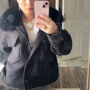 Svart Meotine jacka - Snygg svart jacka från Meotine med stor pälsdetalj på kragen. Jackan har knappar framtill och två fickor med lock på bröstet. Perfekt för kyligare dagar med sin stilrena design och varma känsla. Köpt på plick💓