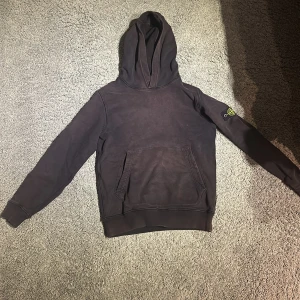 Svart hoodie från Stone Island - Säljer en svart hoodie från Stone Island med klassisk logga på ärmen. Tröjan har en stor ficka framtill och en bekväm huva. Perfekt för en avslappnad stil. Den är rätt solblekt.  