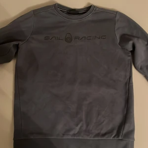 Blå/grå tröja från Sail Racing - Säljer en stilren grå tröja från Sail Racing. Tröjan har en diskret logga på bröstet och är tillverkad i ett mjukt material. Perfekt för en avslappnad look. Passar bra till jeans eller chinos.