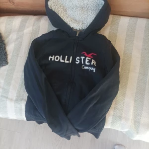 Svart hoodie från Hollister - Mysig svart hoodie från Hollister med vit teddyfoder inuti. Den har en dragkedja framtill och Hollister-logga i vitt och röttpå bröstet. Jätte mysig att ha på sig! 
